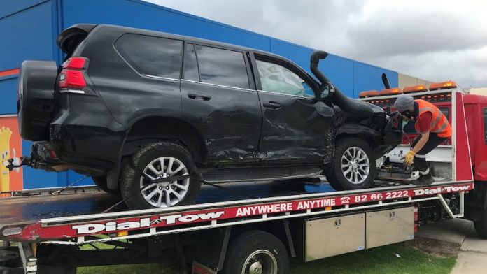 A black 4WD with visible damage to the side sits on a tow truck. A man in an orange vest is securing the car