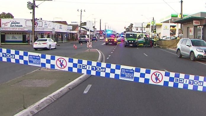 Behind police tape are two ambulances with lights flashing parked on a road with a few other vehicles nearby